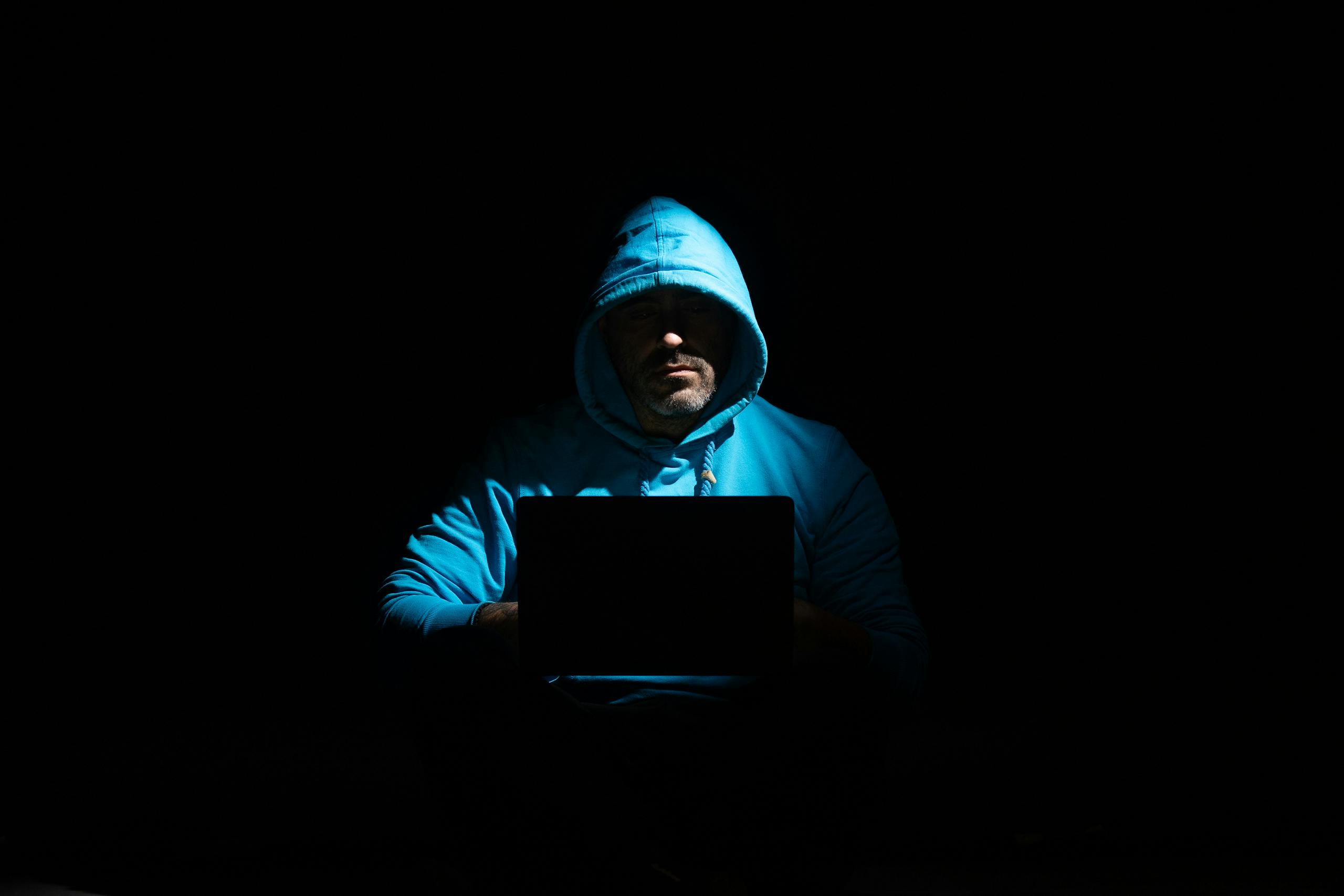 A person in a hoodie using a laptop in a dark setting, conveying mystery and technology themes.