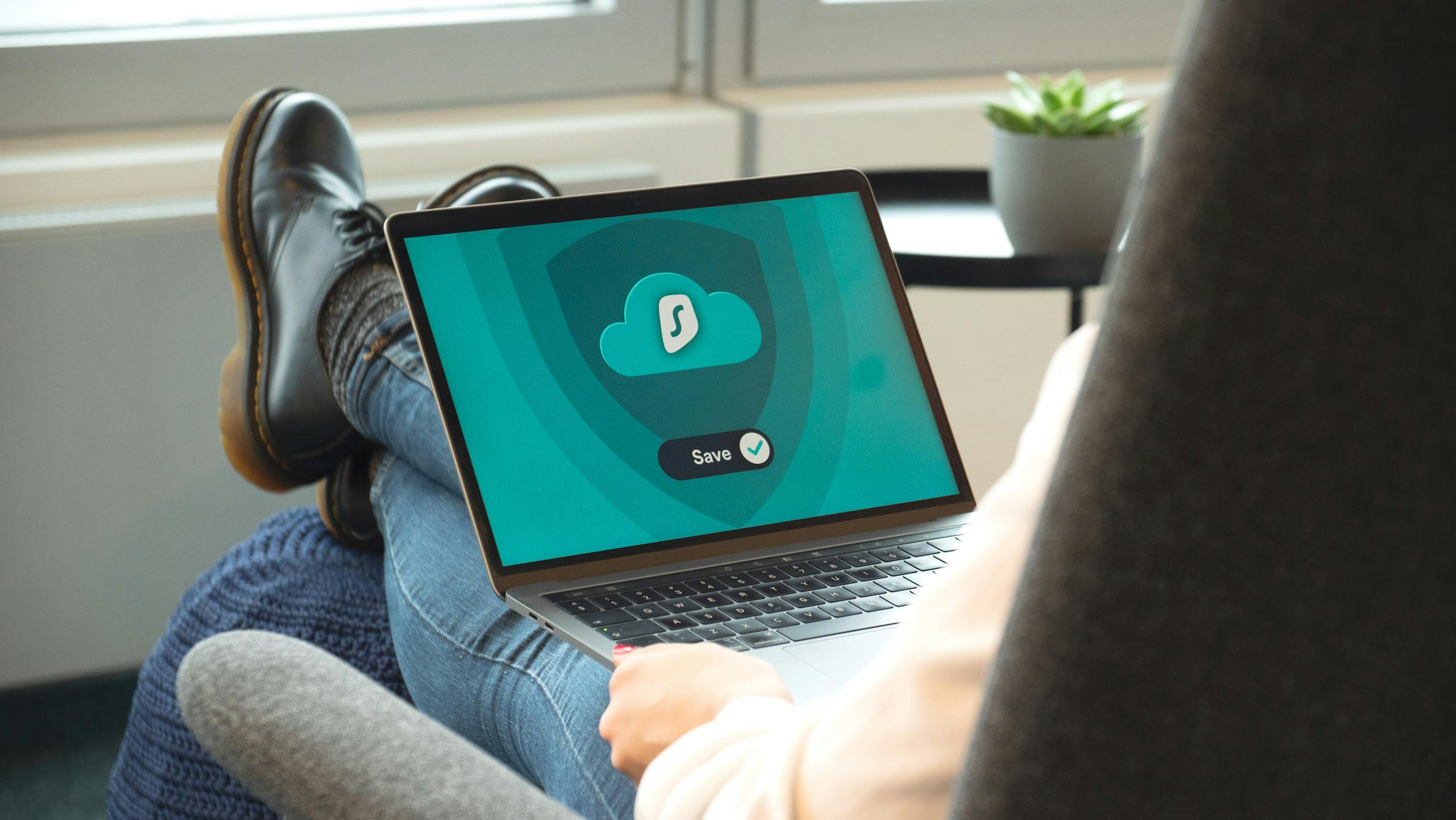 A person wearing boots relaxes with a laptop displaying a VPN save screen indoors.